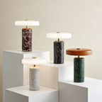 Four modern table lamps with cylindrical marble bases in brown, black, green, and white, paired with round white and brown shades, displayed on white geometric pedestals against a light beige wall.