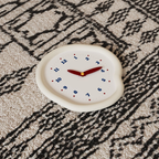 A soft ivory pebble-shaped wall clock with blue numbers and red hands, designed for UK bedrooms, kitchens, hallways and cosy living room walls.