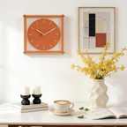 Orange wall clock, yellow flowers in a vase, and a cup of coffee on a table against a white wall with a framed abstract art piece.