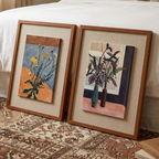 Still Bloom botanical framed wall art set leaning beside a bed with warm wooden frames, earthy floral artwork and cosy bedroom styling.